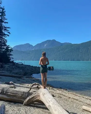Chica de espaldas sobre un tronco en la orilla de un lago rodeado de montañas boscosas