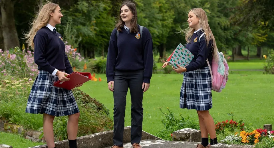 Tre studentesse in uniforme scolastica si trovano nel giardino del Rockwell College davanti a fiori colorati e piante verdi.