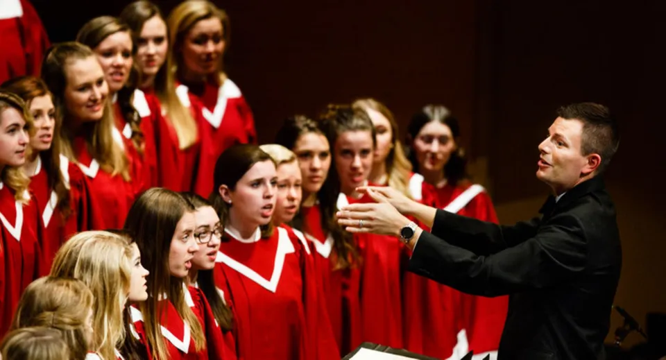 Il coro scolastico della All Saints High School canta in tuniche rosse sotto la direzione di un direttore.