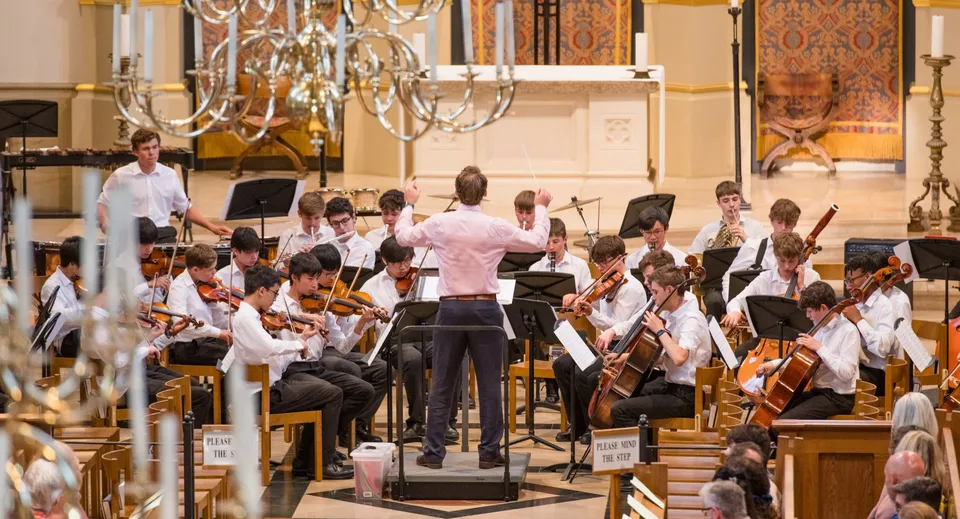 Un'ampia orchestra suona sul palco della Tonbridge School davanti a lampadari decorativi e dettagli architettonici.