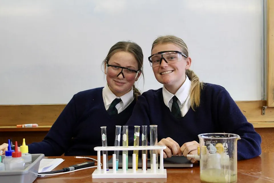 Due studentesse in divisa scolastica mettono in posa sorridenti in un'aula del Columba College.
