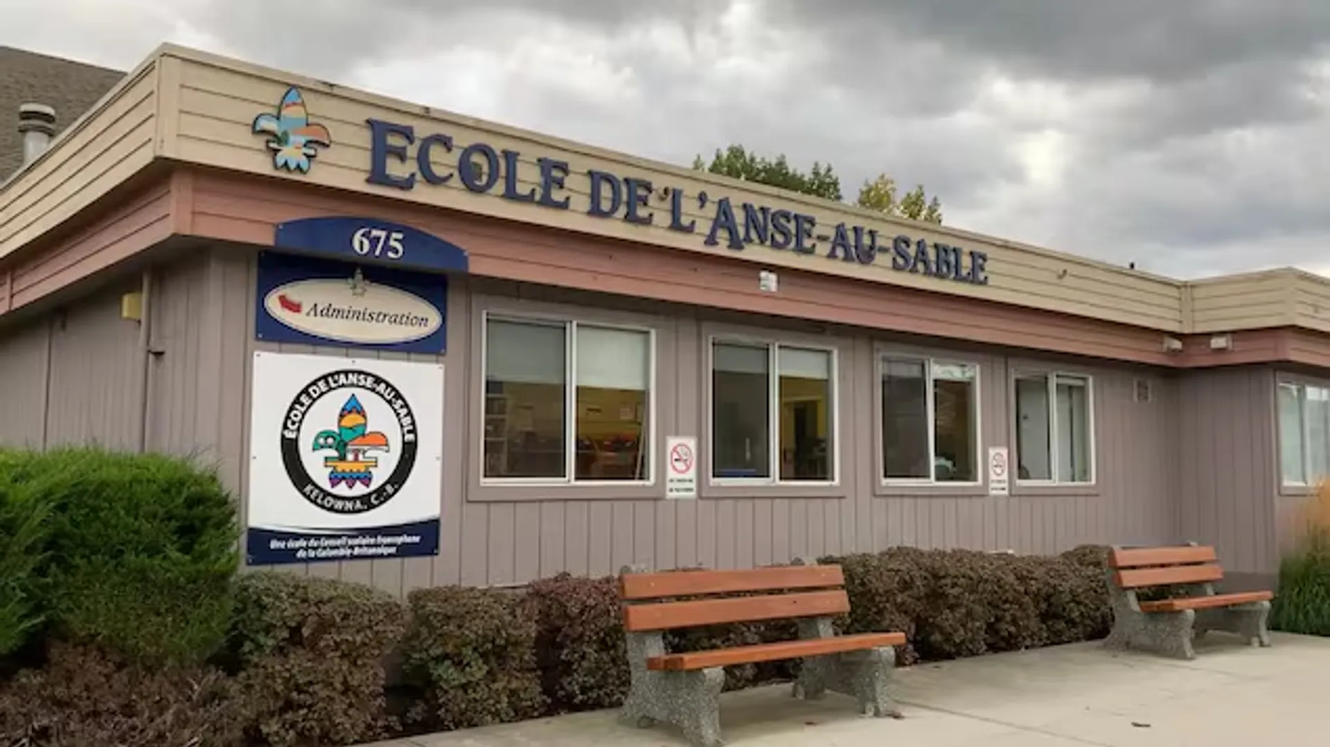 L'edificio a due piani della École de l'Anse-au-sable con segnaletica e panchine in primo piano sotto un cielo nuvoloso.