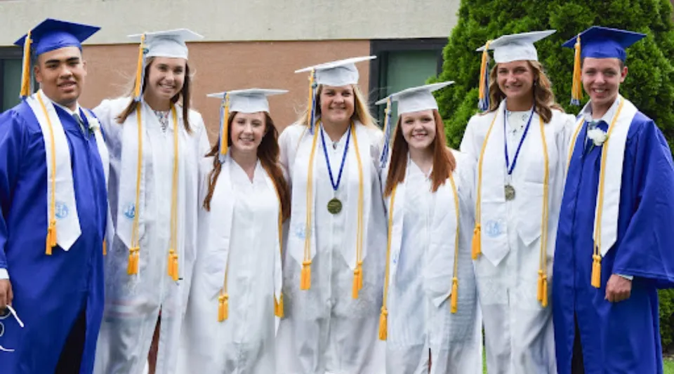 Un gruppo di diplomati della Adrian High School festeggia il diploma in toga blu e bianca davanti a un edificio.