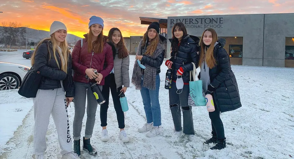 Un gruppo di studentesse si trova all'aperto nella neve davanti alla Riverstone International School con un tramonto colorato sullo sfondo.