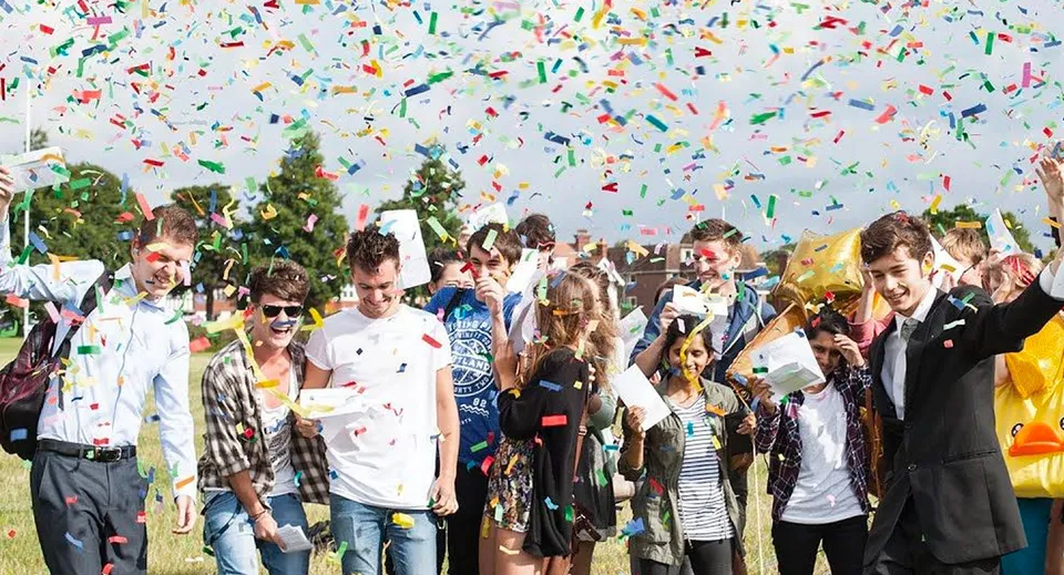 Studenti e studentesse dell'East Sussex College Eastbourne festeggiano all'aperto sotto coriandoli colorati con alberi sullo sfondo.