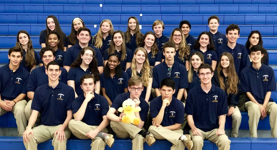 Un gruppo di studenti e studentesse della St. Thomas Aquinas High School posa davanti a una parete blu indossando magliette blu scuro coordinate.