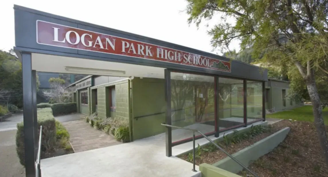 Entrada con el rótulo Logan Park High School ante un edificio escolar verde rodeado de árboles