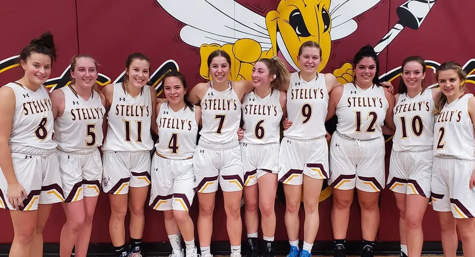 Un gruppo di studentesse della Stellys Secondary School è in piedi con maglie da basket bianche davanti a una parete rossa con una grande figura da cartone animato.