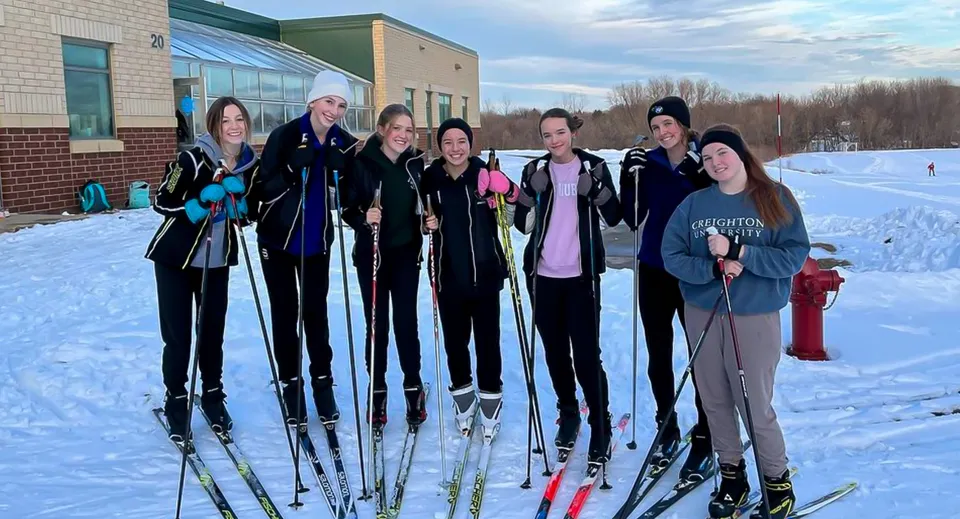 Un gruppo di studenti della Holy Family Catholic High School si trova in abbigliamento invernale sugli sci nella neve davanti a un edificio.