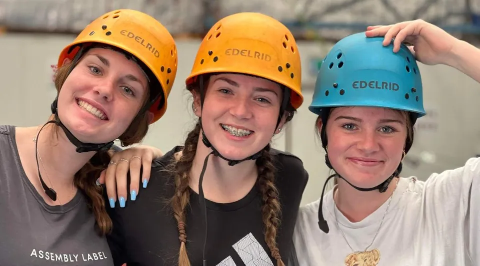Tre studentesse della Carine Senior High School indossano caschi gialli e blu e posano insieme in uno spazio interno.