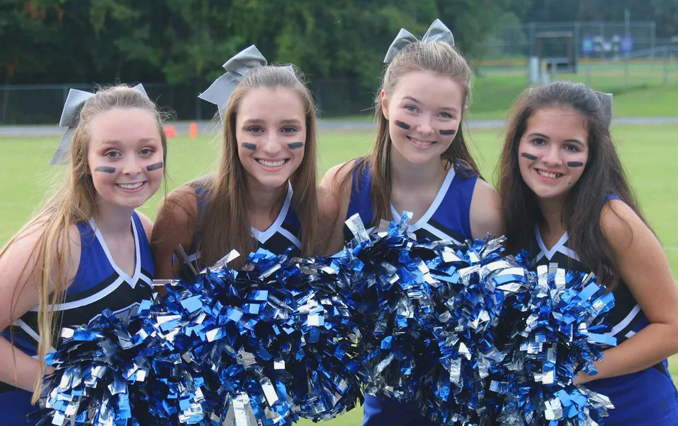 Quattro studentesse della Saint Francis Catholic Academy posano all'aperto come cheerleader con pompon blu e bianchi.