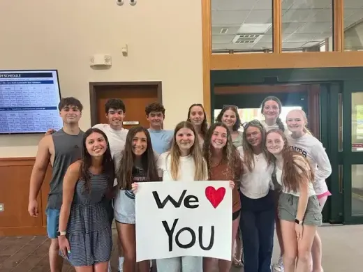 Gruppo di studenti sorridenti con un cartello con scritto We love you in un terminal degli autobus