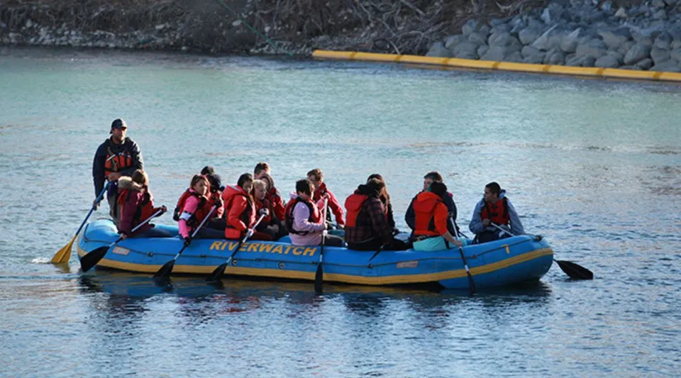 Studenti della William Aberhart High School con giubbotti di salvataggio rossi e arancioni siedono su un gommone blu sull'acqua.