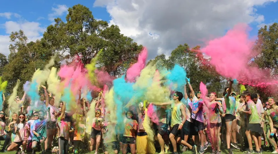 Gli studenti della Thornlie Senior High School festeggiano all'aperto con polveri colorate sotto un cielo blu e una verde vegetazione.