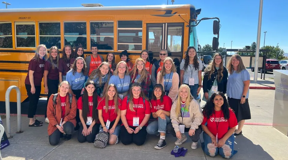 Un gruppo di studentesse dell'Alpine School District davanti a uno scuolabus giallo con il cielo blu sullo sfondo.
