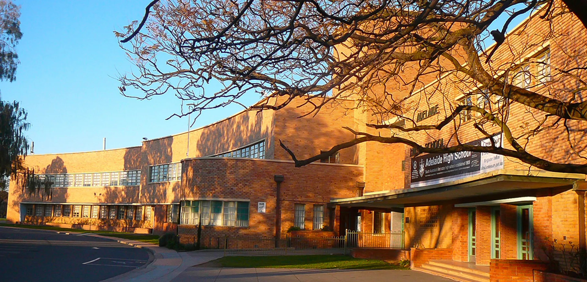 L'edificio in mattoni della Adelaide High School con balcone è circondato da alberi spogli sotto un cielo limpido e blu, bagnato da una calda luce solare.