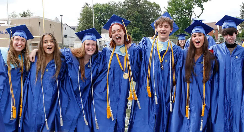 Un gruppo di diplomati del Downingtown Area School District sta insieme all'aperto indossando toge di laurea blu e gialle.