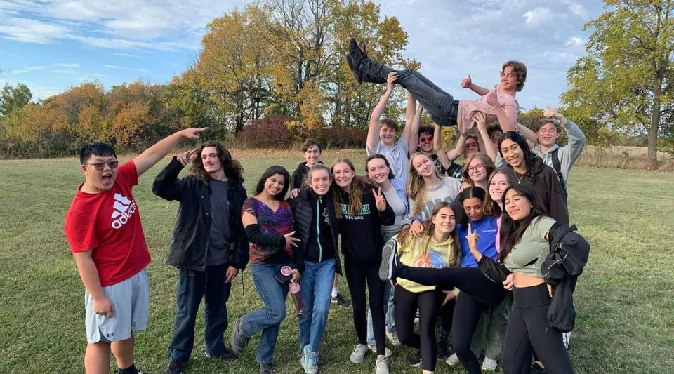 Un gruppo di studenti e studentesse della Thomas A. Blakelock High School si riunisce all'aperto in un'area simile a un parco con alberi e prato.