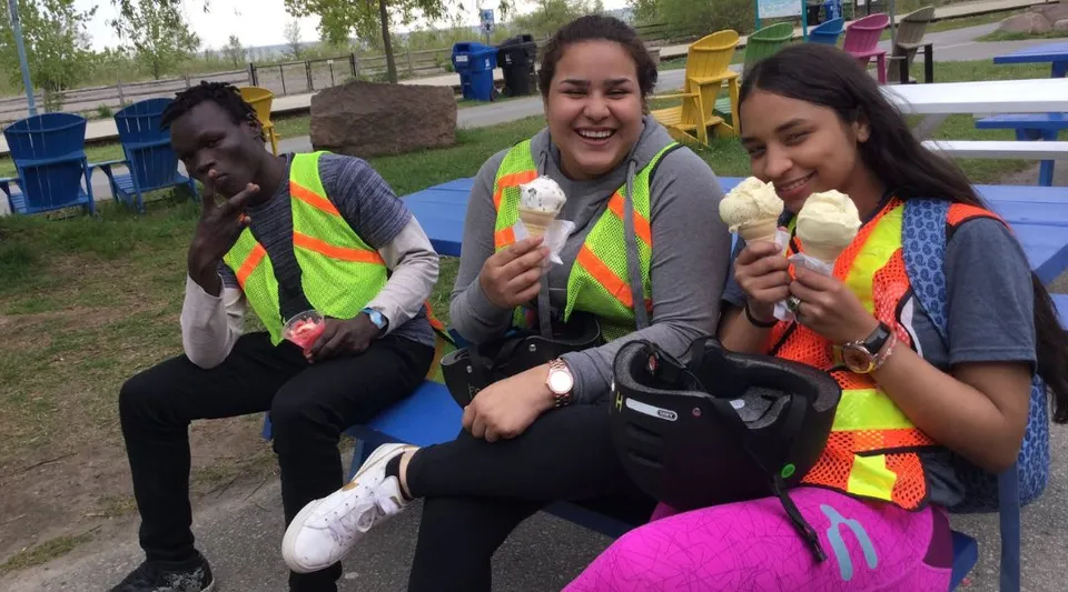 Tre studenti della Greenwood Secondary School siedono nel cortile della scuola e mangiano un gelato davanti a un parco giochi colorato.