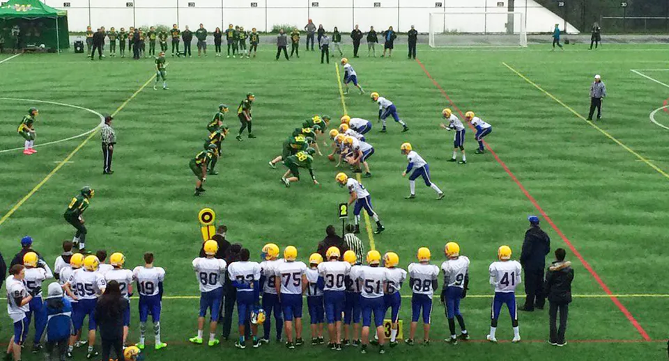 Un gruppo di giocatori di calcio si trova sul campo da gioco della Windsor Secondary School con spettatori e allenatori sullo sfondo.