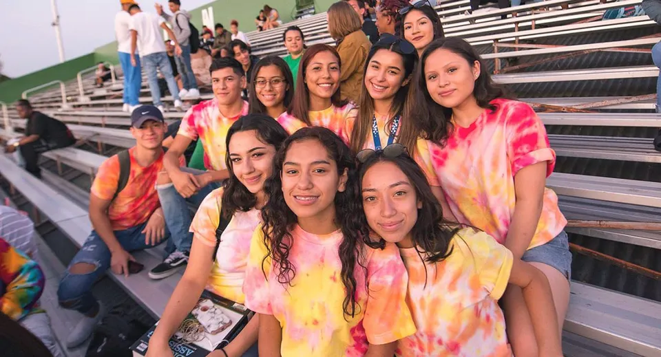 Un gruppo di studentesse sorridenti in magliette tie-dye colorate siede sulle tribune del campus dell'Antelope Valley Union High School District con vista sul campo sportivo.