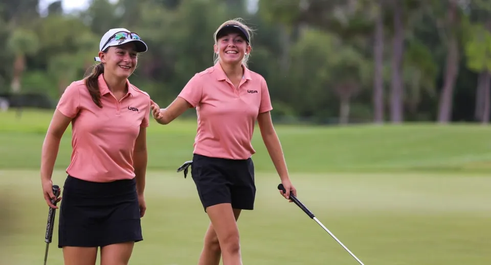 Due giovani golfiste della Bishop’s Gate Golf Academy camminano sorridenti con le mazze su un campo da golf.