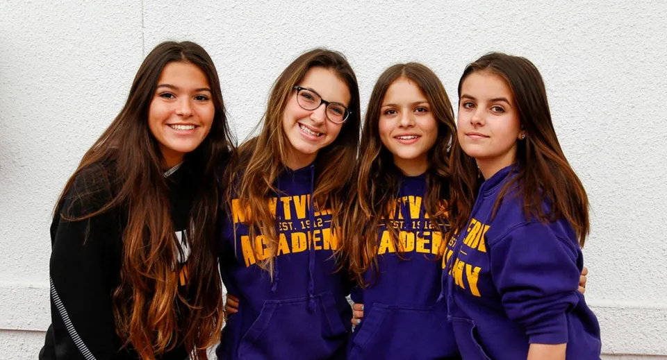 Quattro studentesse della Montverde Academy posano sorridenti davanti a una parete bianca con felpe viola identiche.