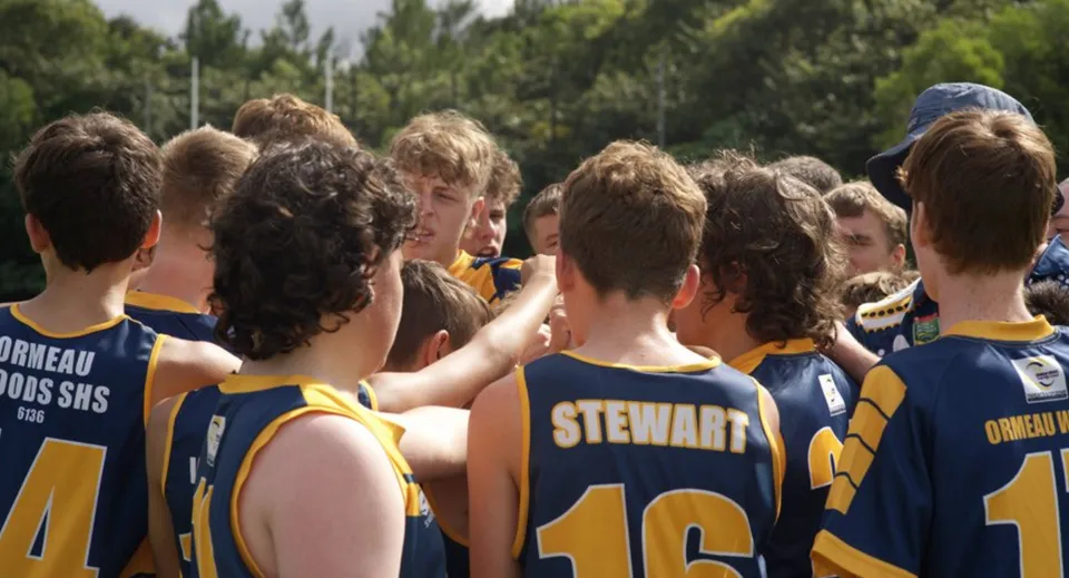 Un gruppo di giovani atleti si trova all'aperto su un prato nel campus della Ormeau Woods State High School con vista sugli alberi.