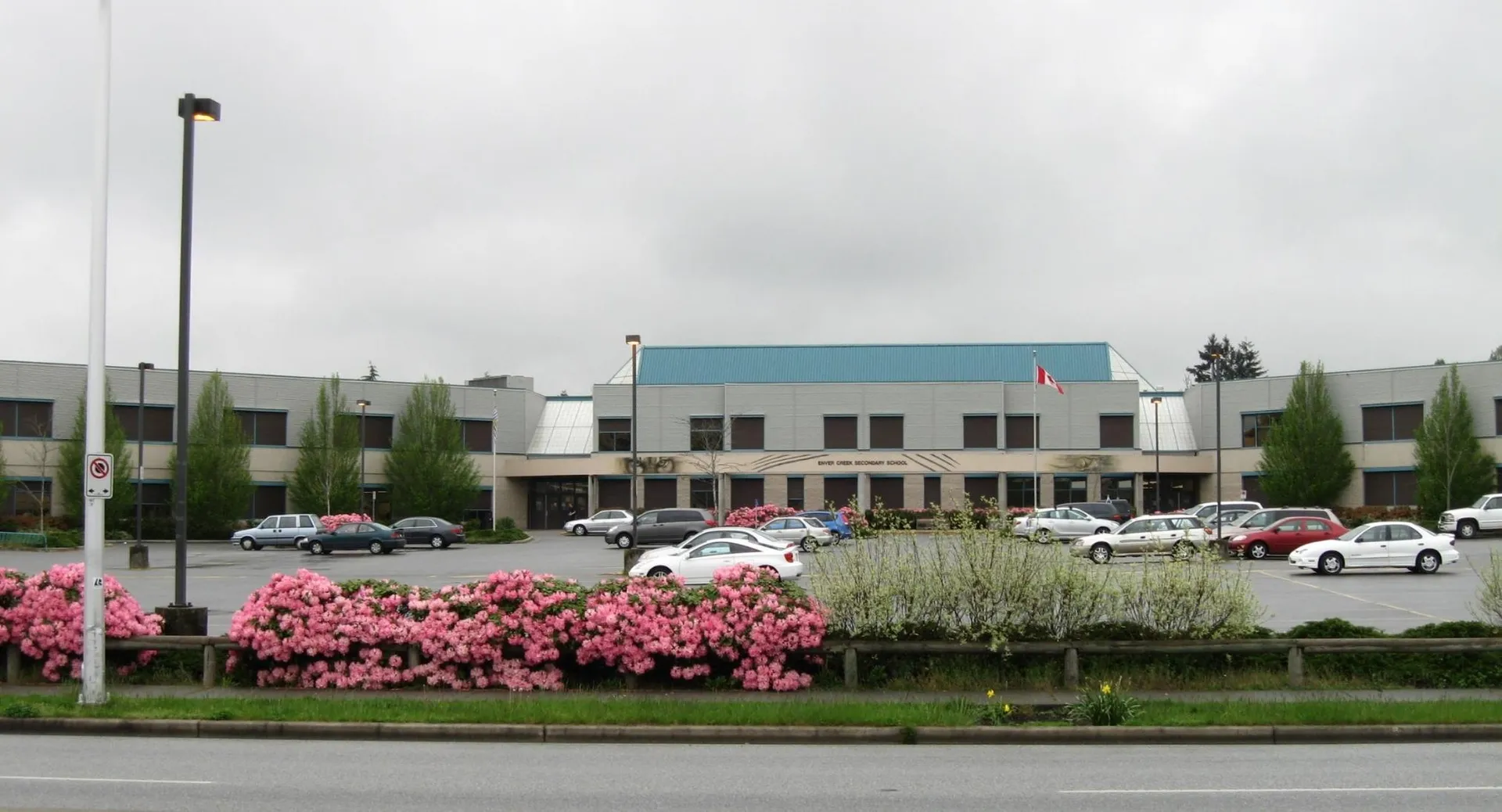 L'edificio della Enver Creek Secondary School con un parcheggio in primo piano e fiori rosa in fiore sotto un cielo nuvoloso.