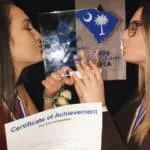 Dos chicas jóvenes besan un trofeo transparente mientras sostienen un certificado de logro