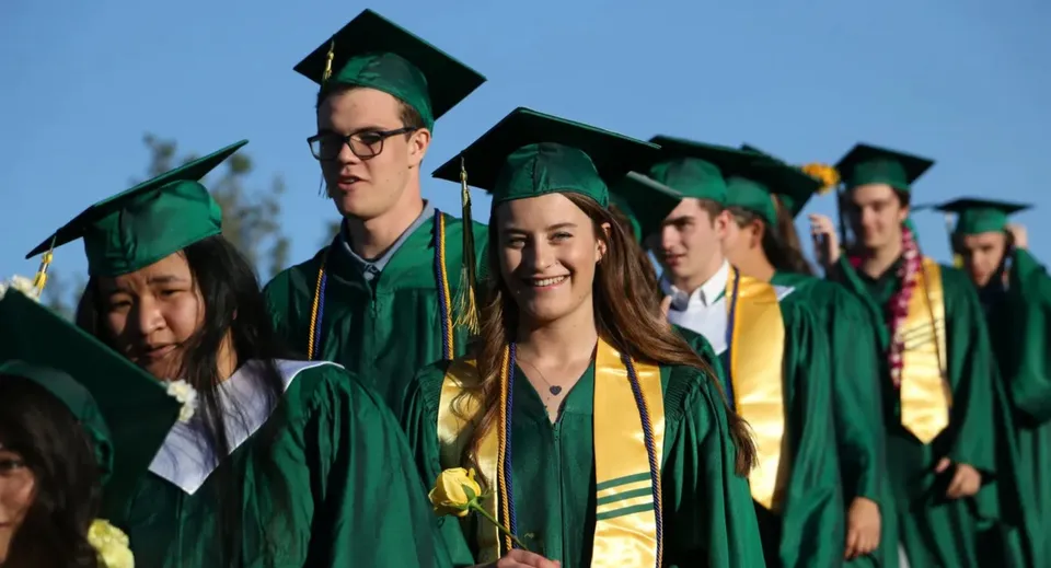 Un gruppo di diplomati della St. Joseph Academy indossa toghe verdi e gialle davanti a un cielo azzurro e limpido.
