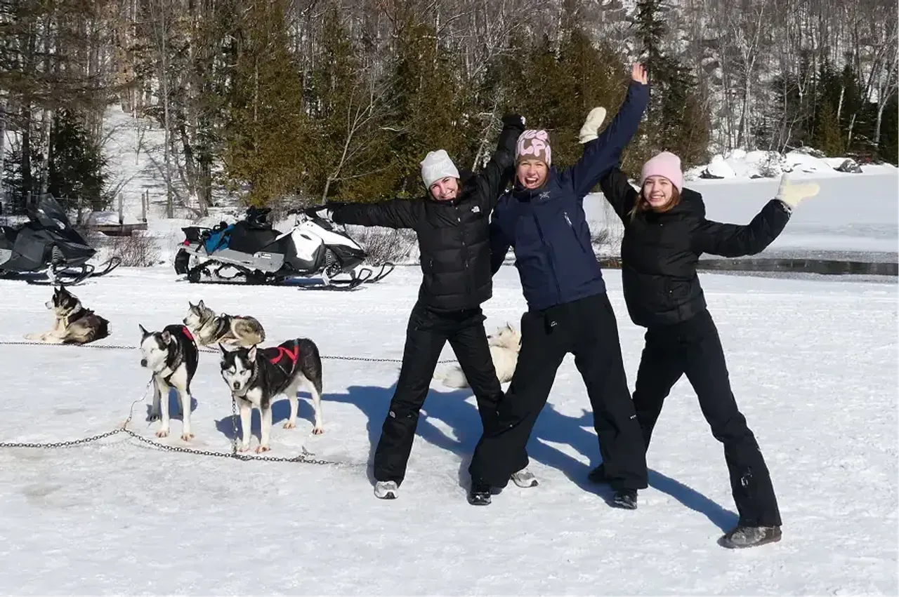 Tre studentesse esultano nella neve accanto a cani da slitta husky durante un anno all'estero in Canada