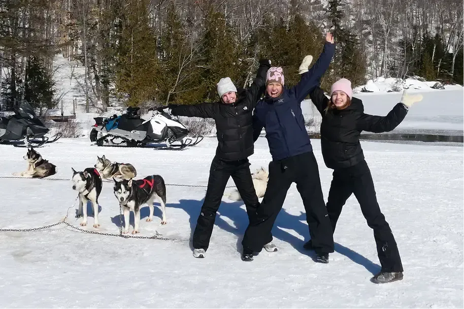 Tre studentesse esultano nella neve accanto a cani da slitta husky durante un anno all'estero in Canada