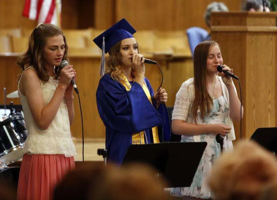 Tre studentesse della Immanuel Christian Secondary School cantano con la toga da diploma su un palco con microfoni.