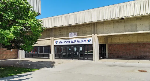 Das Gebäude der W.P. Wagner High School mit einem Willkommensschild über dem Eingang ist von Bäumen und Grünflächen umgeben.
