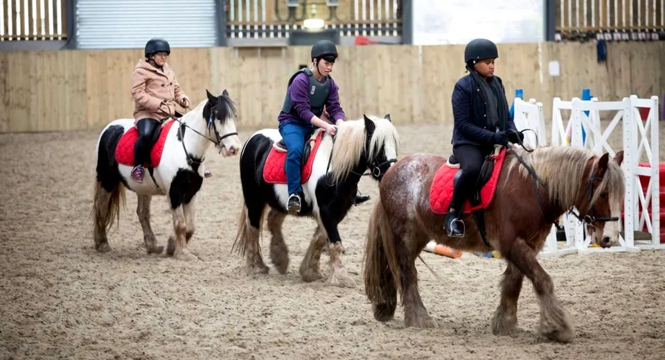 Tre persone in tenuta da equitazione partecipano a una lezione di equitazione della Worthgate School Canterbury in un maneggio coperto.