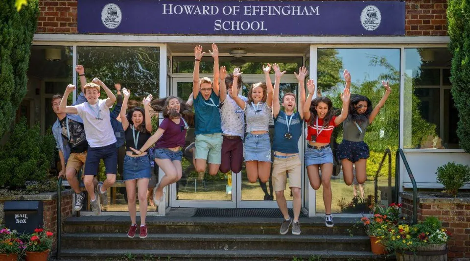 Un gruppo di studenti e studentesse è in piedi con le braccia alzate davanti alla Howard of Effingham School e festeggia.