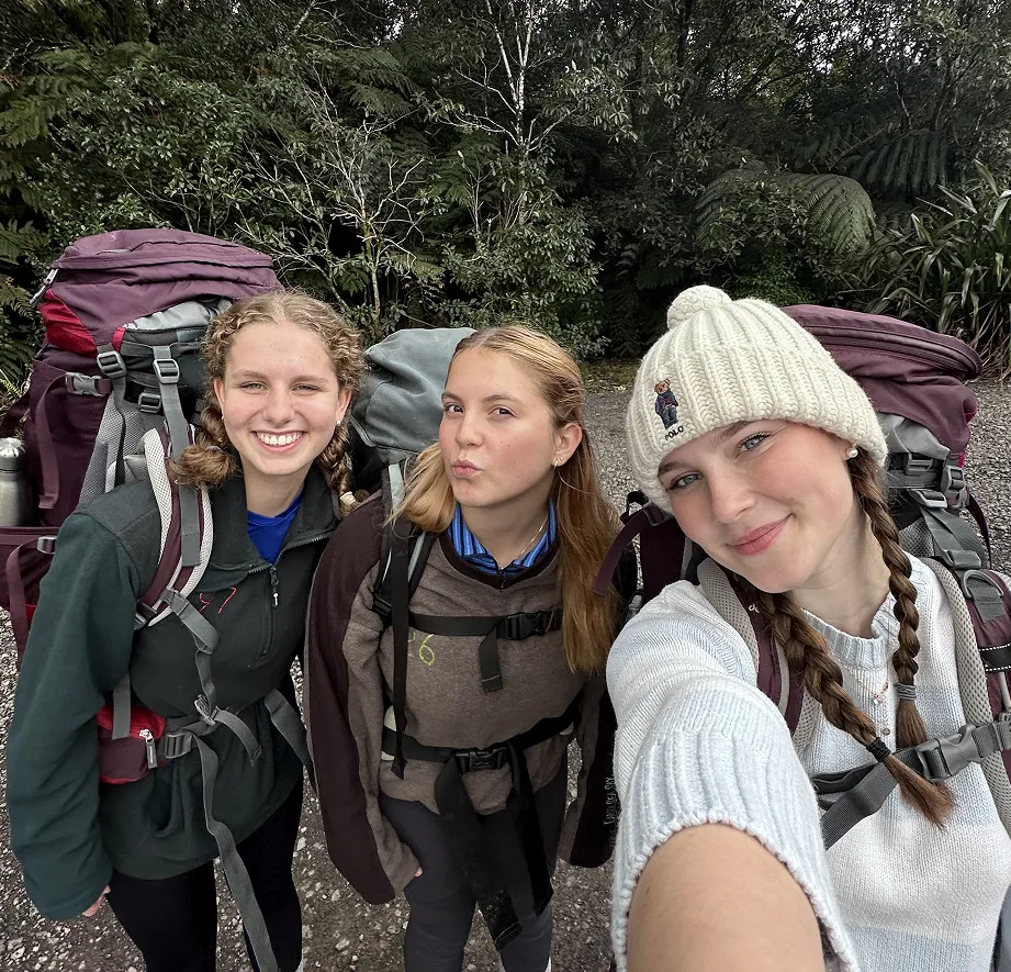 Tres estudiantes con grandes mochilas de trekking sonríen en un selfie en el bosque