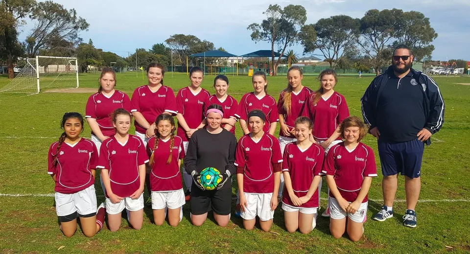 Un gruppo di giovani calciatrici in maglia rossa posa con un allenatore su un campo in erba al Seaford Secondary College.