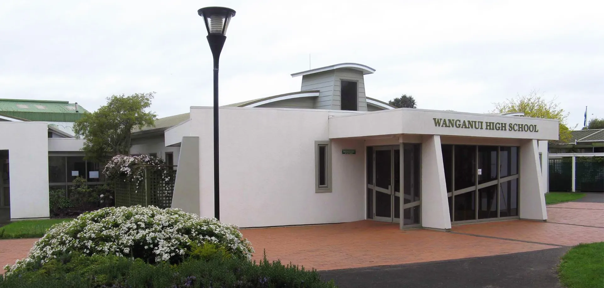 L'edificio scolastico moderno e bianco della Whanganui High School con l'entrata principale e un alto lampione in primo piano, circondato da verde e fiori.