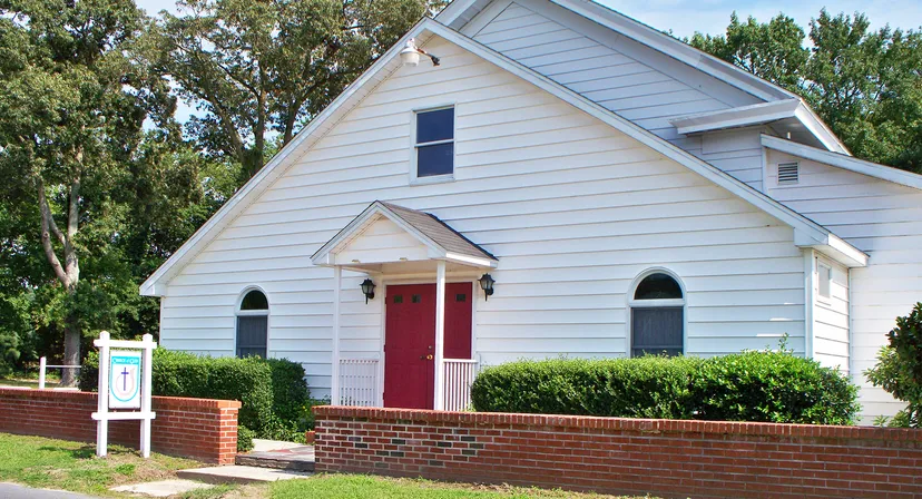 L'edificio bianco della chiesa con porta rossa e finestre ad arco, situato nel campus della Beacon of Hope Christian School, è circondato da alberi verdi.