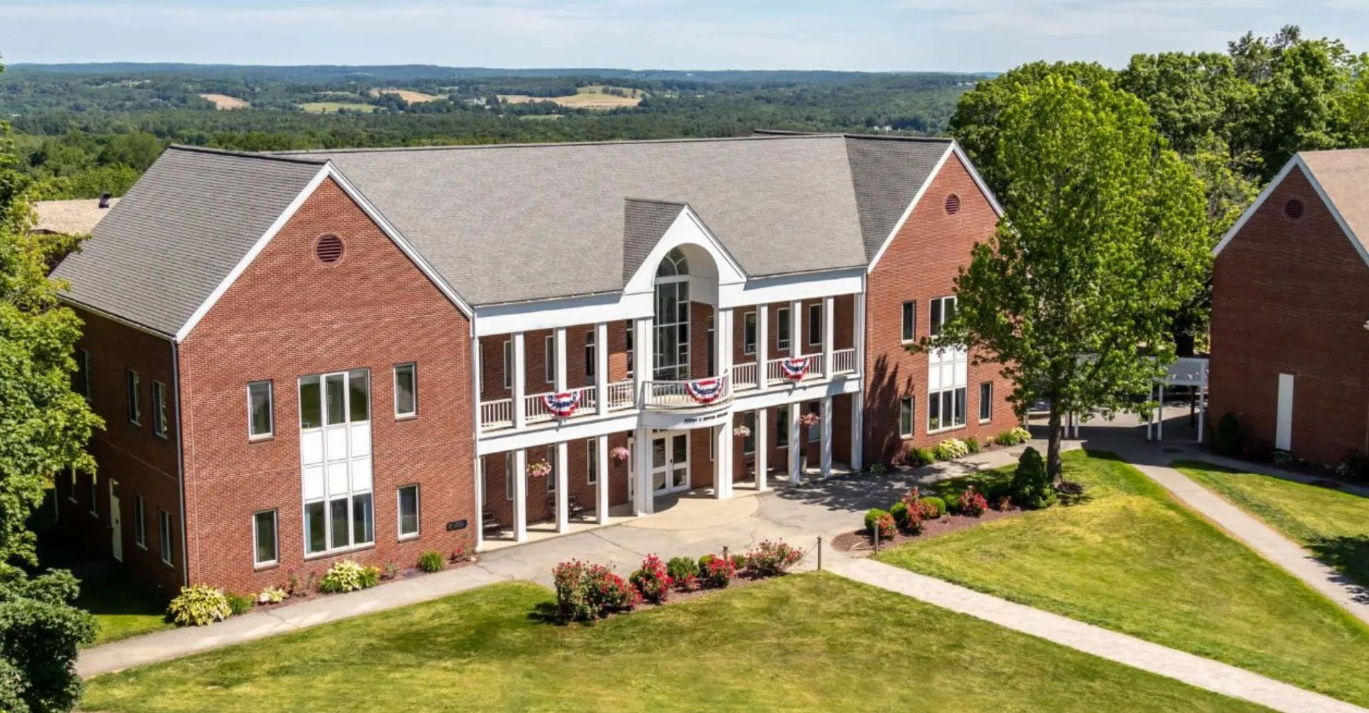 L'edificio in mattoni a più piani della Woodstock Academy si erge davanti a colline verdi e circondato da una lussureggiante vegetazione.