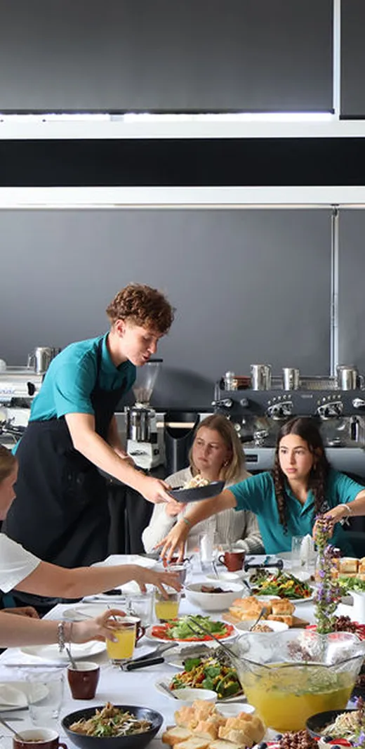 Estudiantes sirven platos y ensaladas en una larga mesa durante una clase de cocina