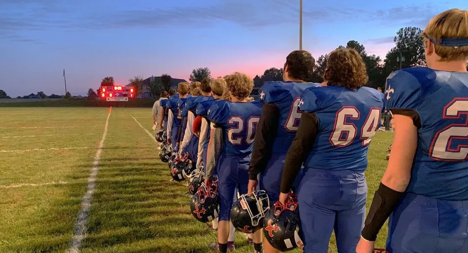 Un gruppo di giocatori di calcio in maglia blu si trova sul campo da gioco della Saint Paul Lutheran High School al tramonto.
