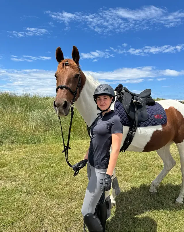 Studentessa accanto a un cavallo pezzato sellato in un prato, pronta per l'equitazione