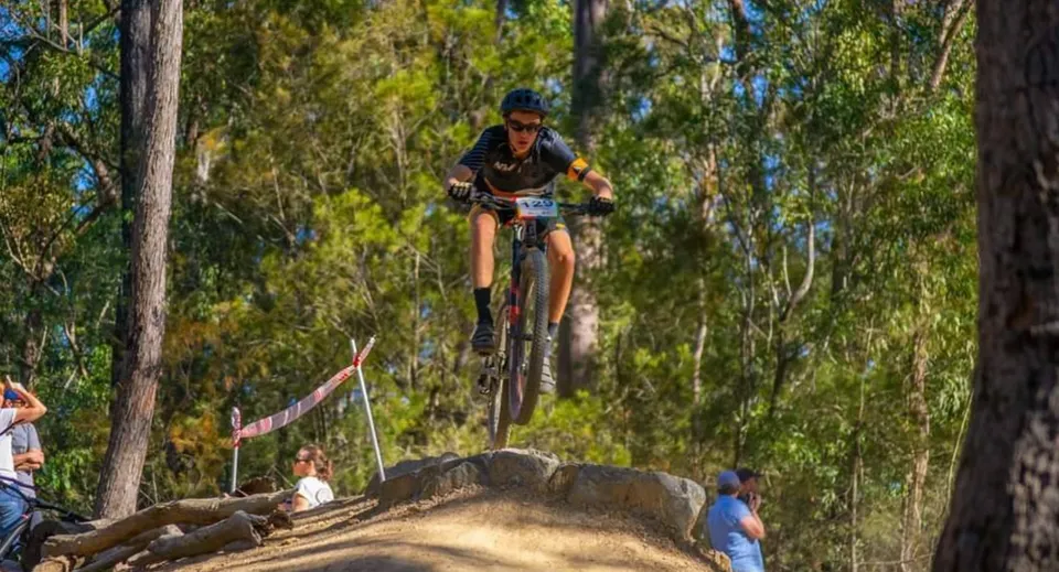 Un mountain biker esegue un salto su una rampa di terra nel terreno della Alexandra Hills State High School, mentre gli spettatori guardano.