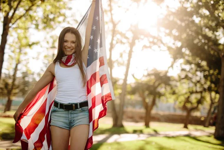 Lächelnde junge Frau hält eine amerikanische Flagge in einem sonnigen Park