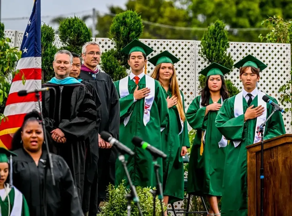 Neodiplomate e neodiplomati della Torrance South High School in toga verde e nera sono all'aperto davanti a una bandiera americana.