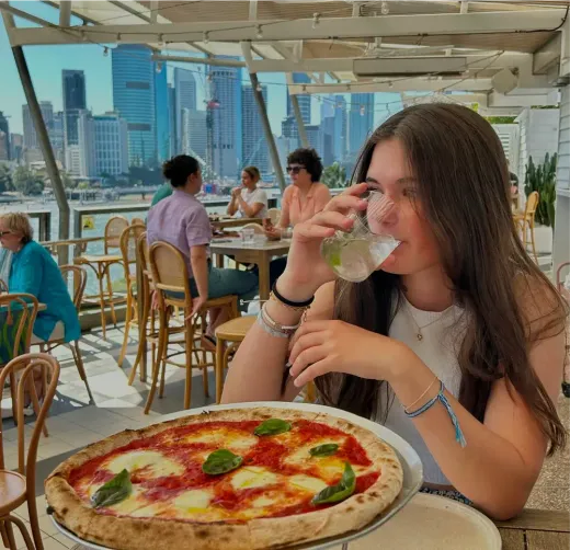 Luisa trinkt aus einem Glas in einem Außenrestaurant mit Pizza und Stadt-Skyline in Australien
