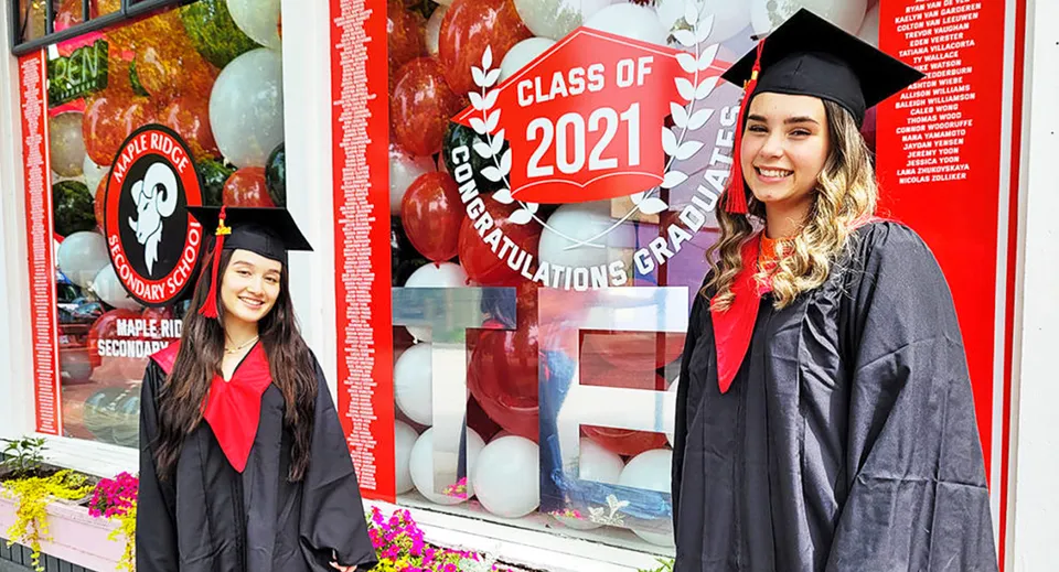 Due studentesse in toga da diploma stanno davanti a una decorazione della Class of 2021 nel campus della Maple Ridge Secondary School.
